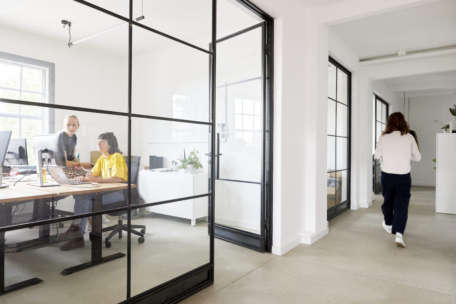 Blick in ein Büro wo zwei Personen arbeiten, eine weitere Person läuft über Flur